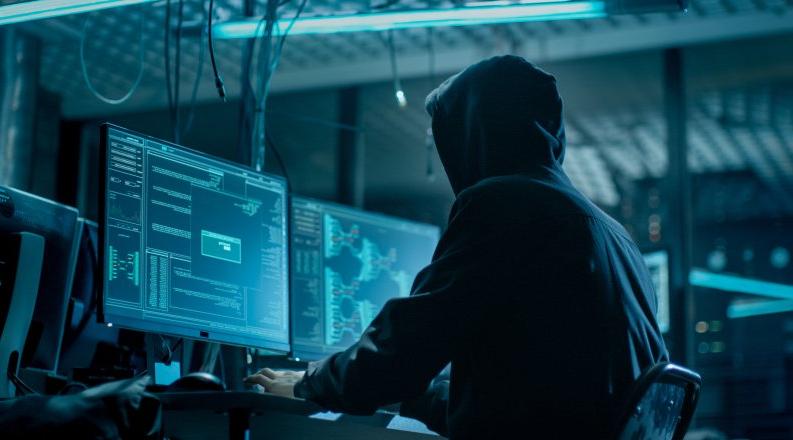 Shot from the Back to Hooded Hacker Breaking into Corporate Data Servers from His Underground Hideout. Place Has Dark Atmosphere, Multiple Displays, Cables Everywhere.